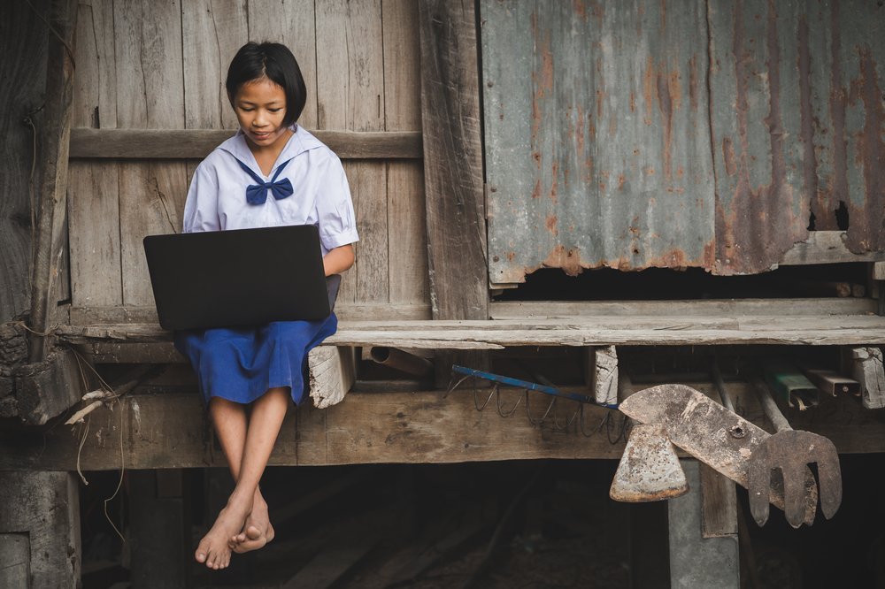 A girl in a blue school uniform sits on the front step of a raised building made of wood and corrugated iron. She is typing on a laptop.