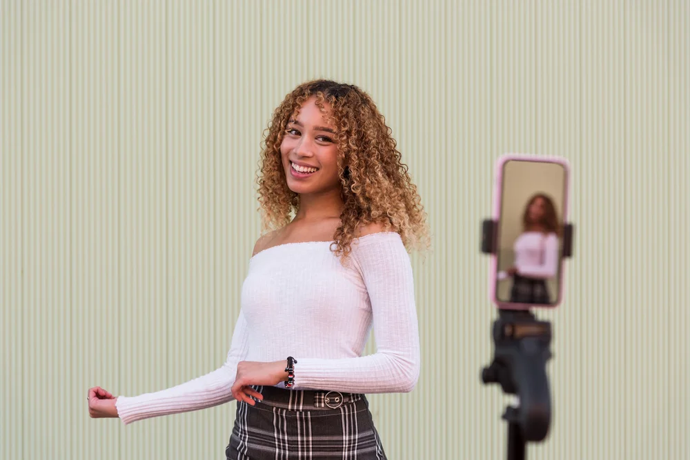 A young woman films a video with a phone on a tripod.