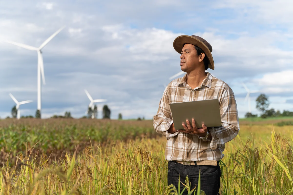A person standing outside in a field, holding a laptop,  with wind turbines behind them.