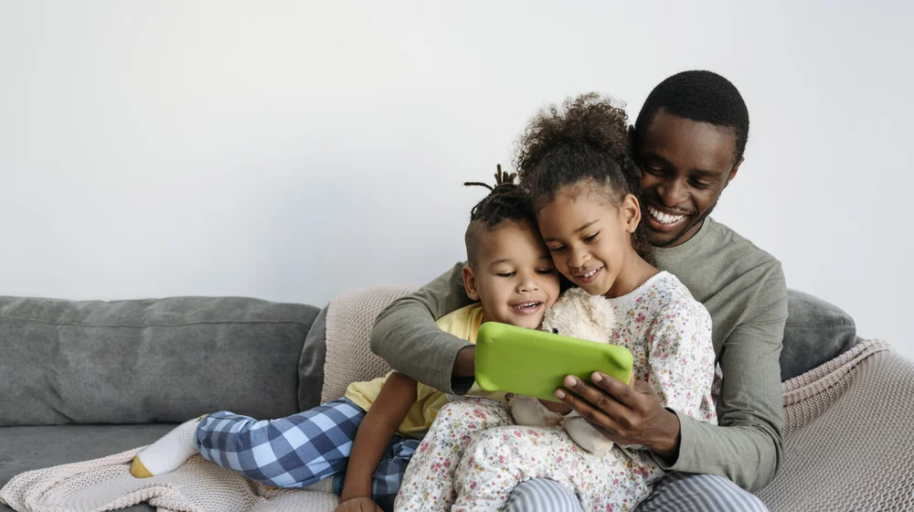 A smiling man with son and daughter are using a digital tablet on a sofa