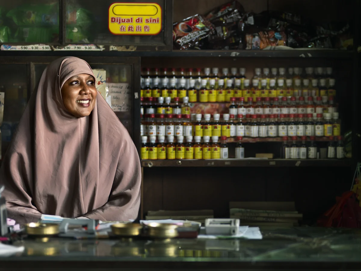 A woman in a dusk-pink hijab behind the counter at her store, smiling at somebody out of shot. There are bottles with bright yellow and white labels on shelves in the background.