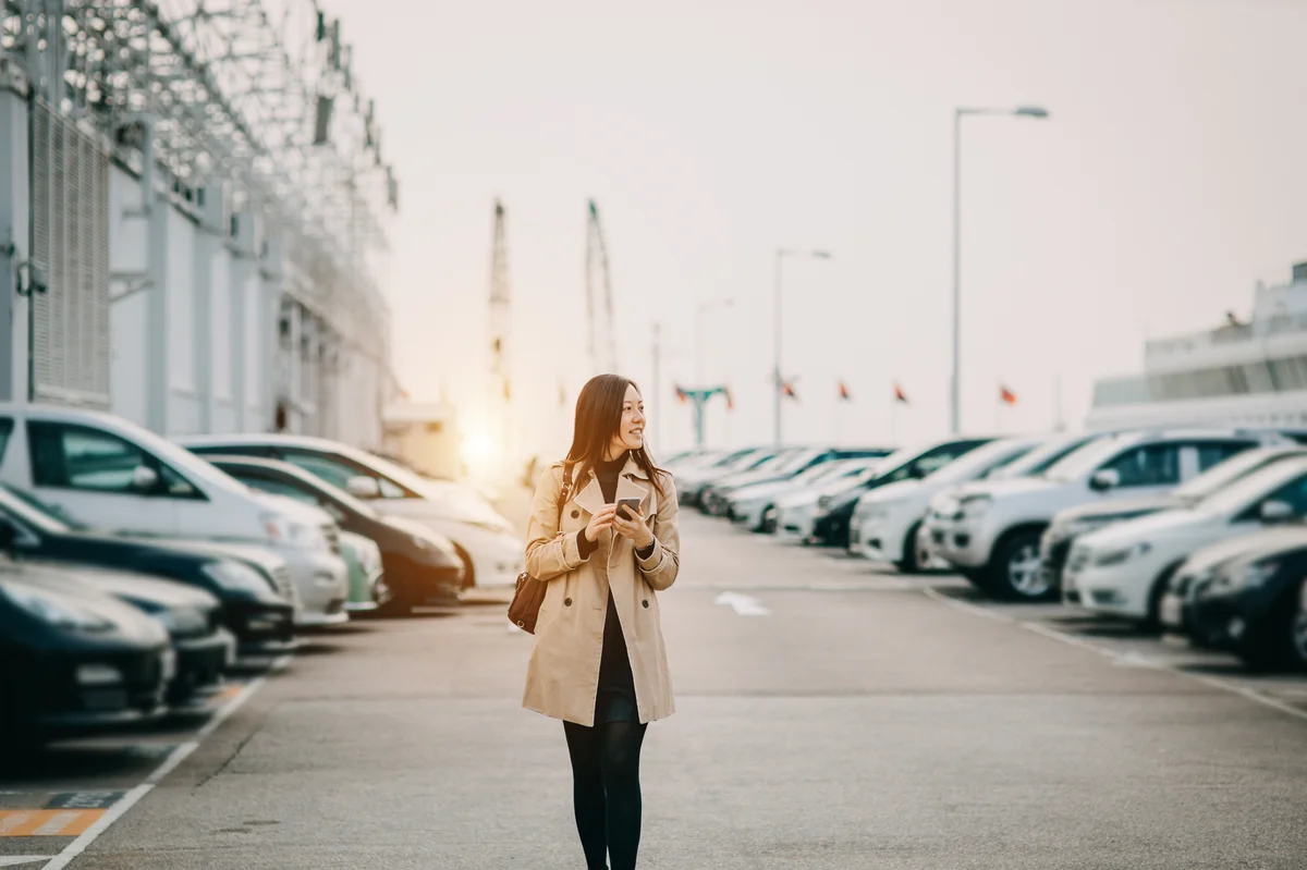 A woman holding her smartphone looks around at an outdoor car dealership.