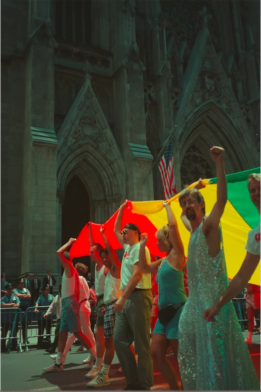 A group of people marching along a street carrying a colorful flag over their heads