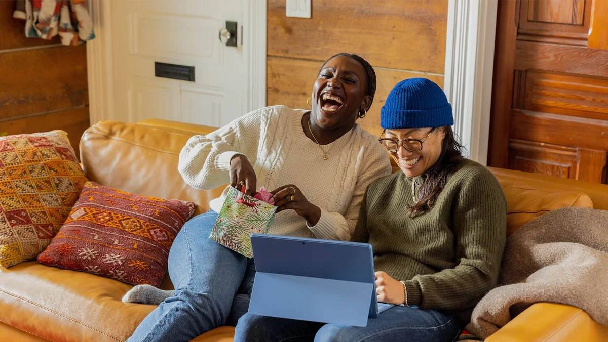 Two women sitting on the couch looking at a tablet. Both women are laughing.