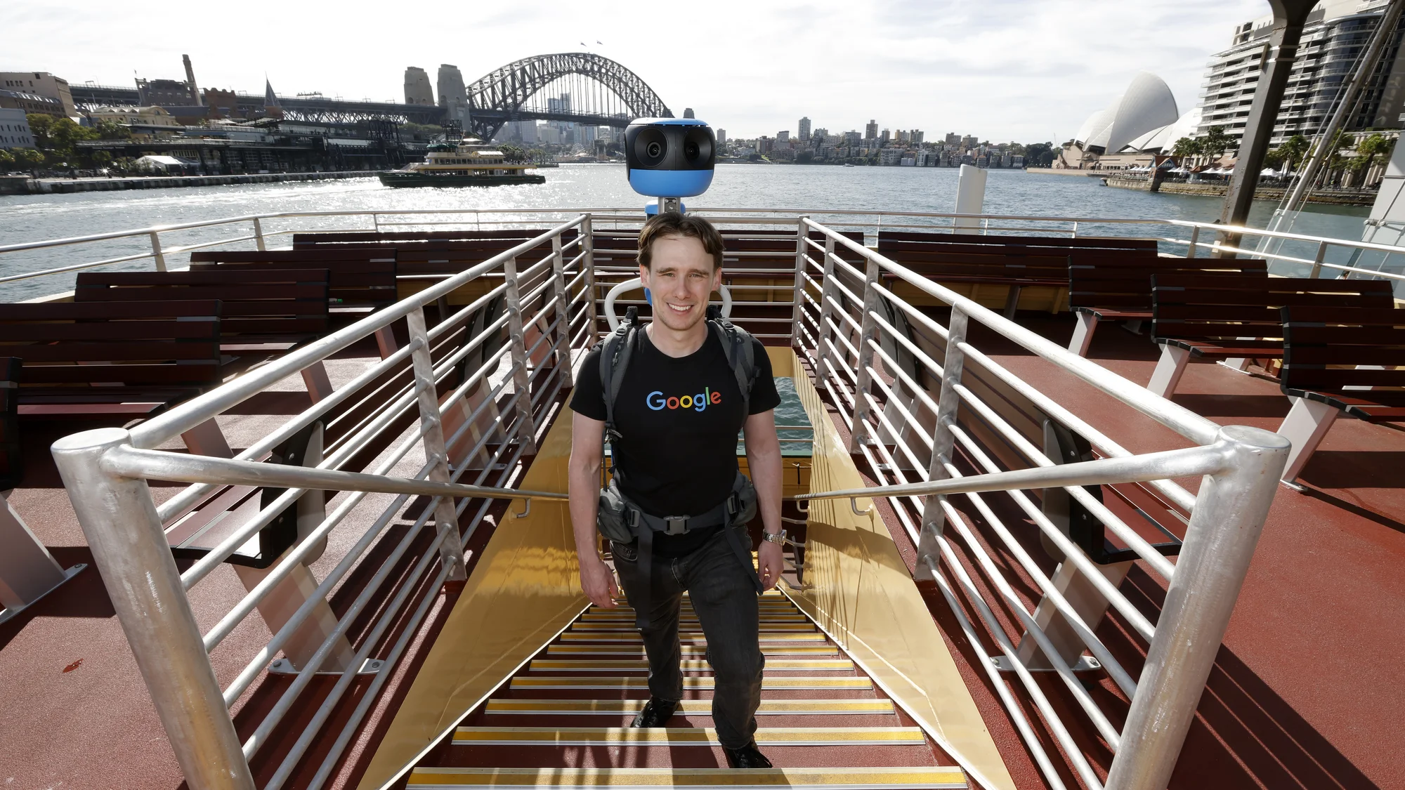 A photo showing Daniel Nadasi walking up the stairs on the outside deck of a green and yellow ferry wearing the blue Street View trekker backpack.