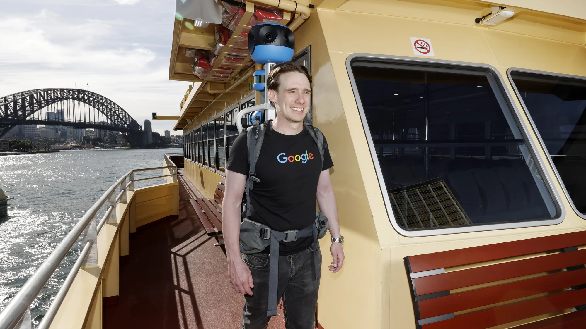 A photo showing Daniel Nadasi walking down the side of the outside deck in a green and yellow ferry, wearing the blue Street View trekker backpack.