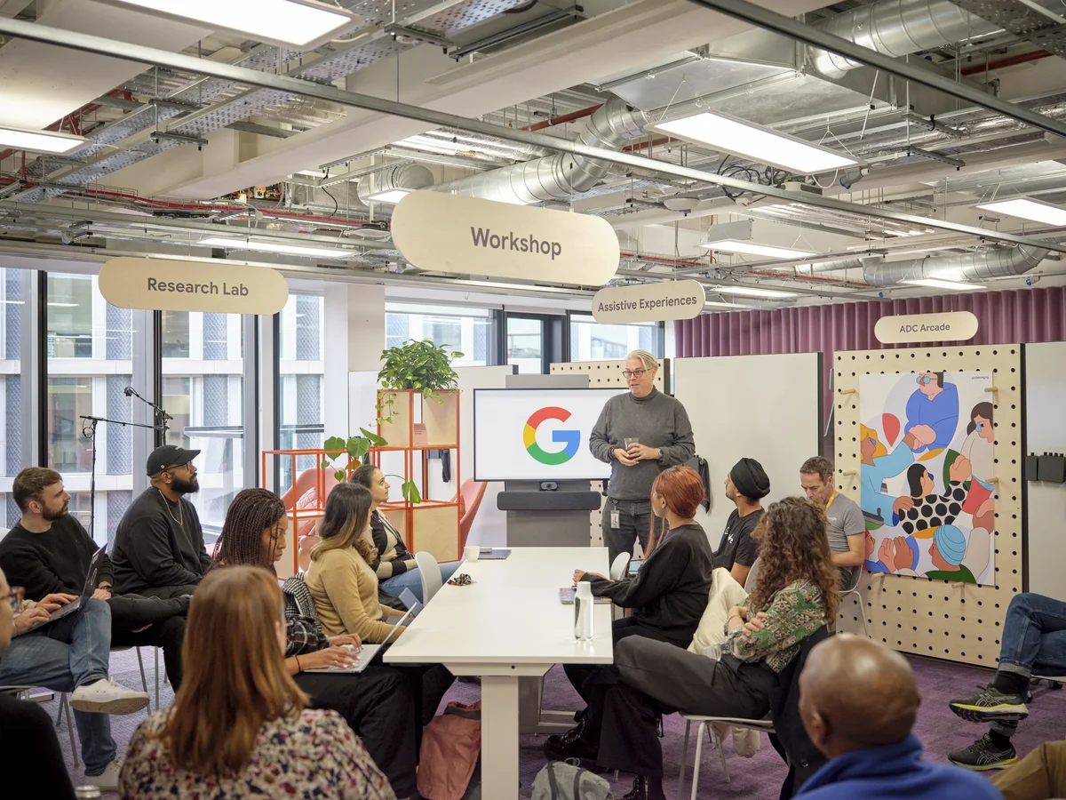 A group of about twelve people are sitting around a white table in the Accessibility Discovery Centre, listening to a man standing in front of the group holding a drinking glass and standing next to a screen with a Google “G” logo on it. From the ceiling