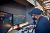 A man wearing a cap is pictured in the ADC Arcade. He is looking at two screens and using a chin-controlled joystick and some multi-coloured buttons to play a video game.