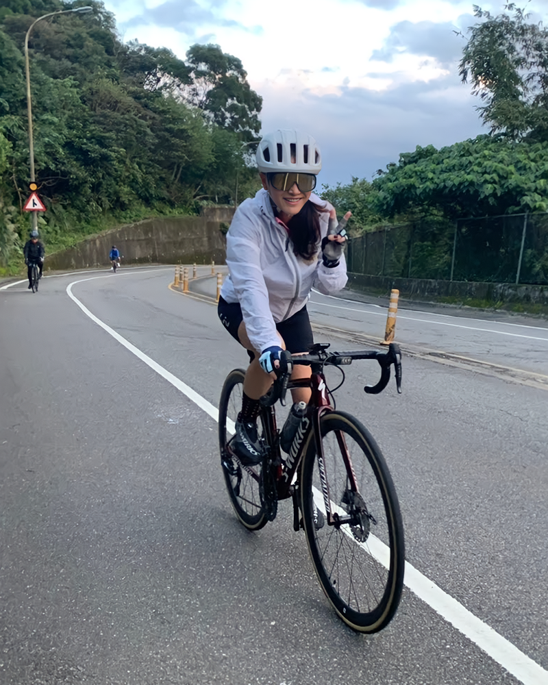 Frances is smiling as she rides a bicycle on a road. There are lush trees next to her.