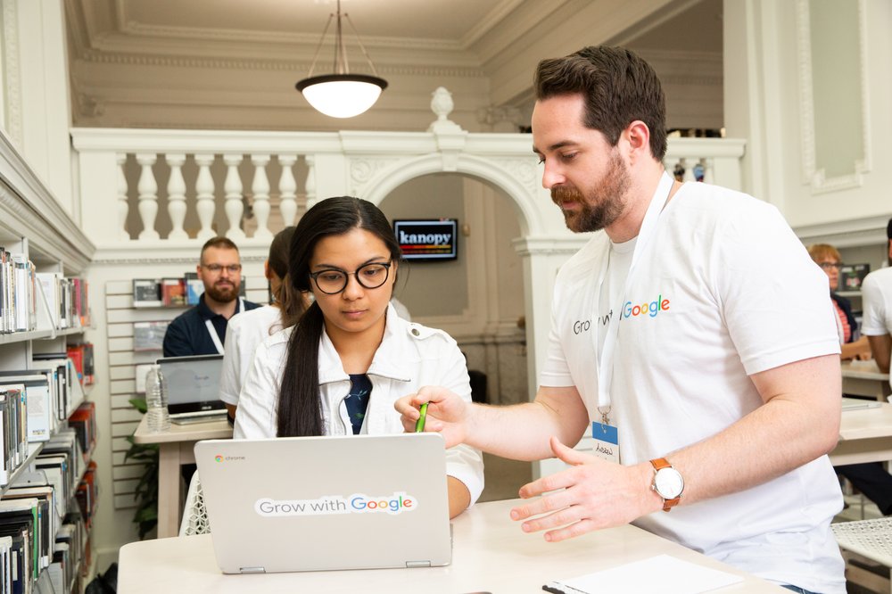 A woman sits at a laptop while a man in a Grow with Google shirt stands up and talks to her