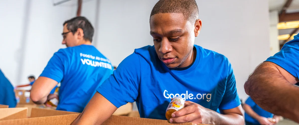 A Google employee in a blue Google.org t-shirt loads a jar of food into a cardboard box for delivery. Other volunteers in the same kind of blue t-shirt can be seen in the background, in a warehouse setting.