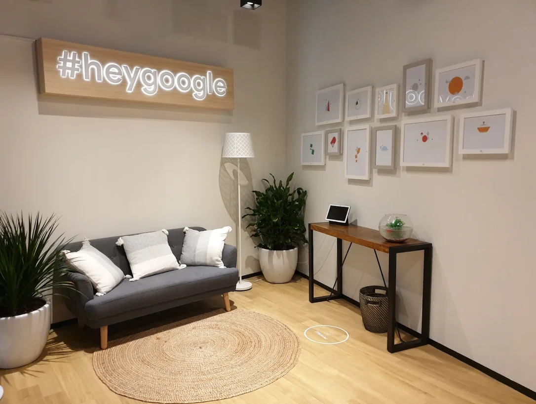 A room with a couch, a table, a tablet and different kinds of lights and decor placed around the room. One wall is decorated with a “#heygoogle” sign.