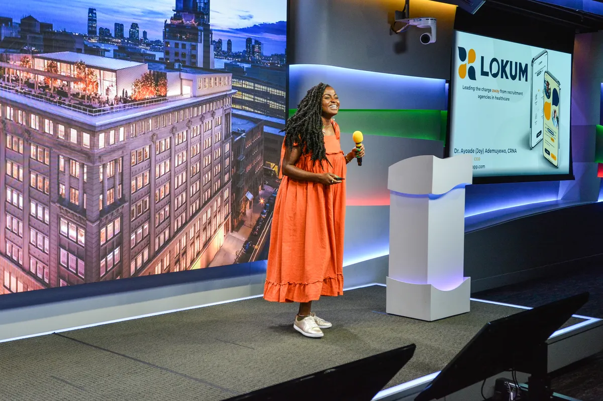 Joy Ademuyewo stands center stage in an orange dress holding a microphone as she addresses an audience in New York City. The screens behind her show a building on one side and the Lokum logo on the other.