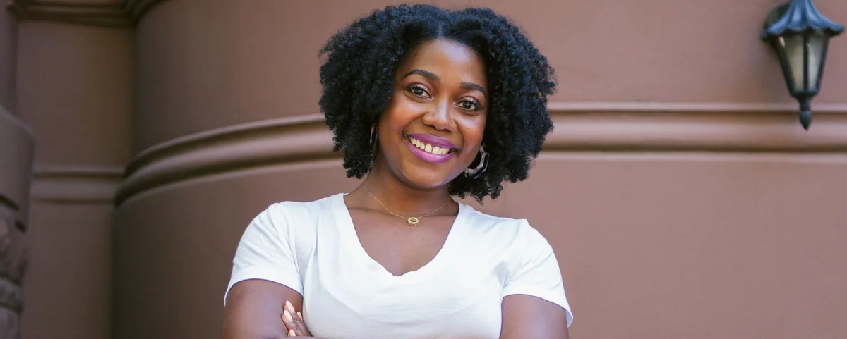 A Black woman wearing a white shirt smiles at camera while standing in front of a classic NYC brownstone building.