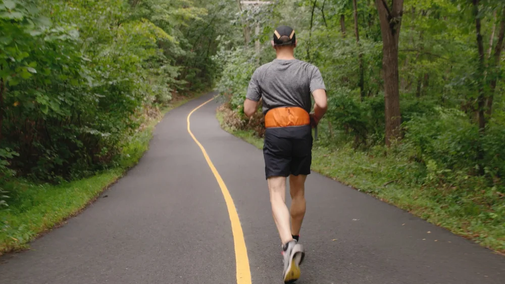 Image shows curved road with tall green trees on either side. A man is in the middle wearing running shorts and t-shirt with his back to the camera and is mid-stride. He is running alongside a yellow line that has been painted in the middle of the road.