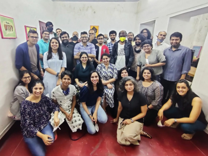 This picture shows The News Minute team. There are a group of people inside an office room. Some of the group are seated on the floor while others are standing behind them. There are 28 people visible. The room has white walls and the floor is a red color.