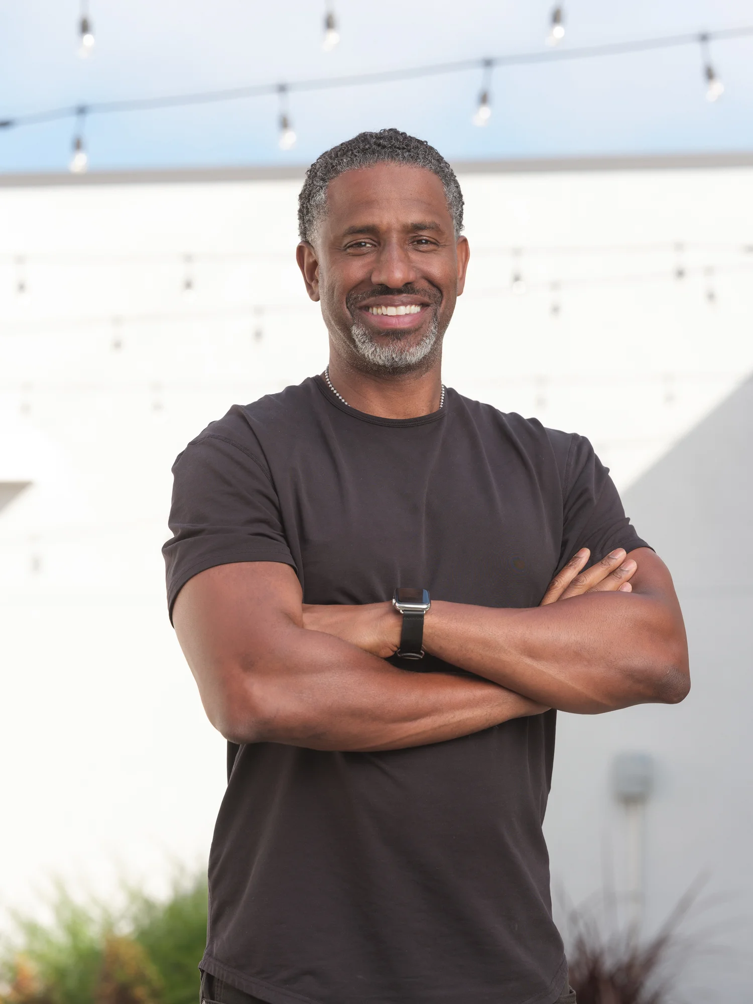 Hamet Watt folds his arms and smiles into the camera. There is a white wall behind him, as well as some hanging lights. He is wearing a black t-shirt.
