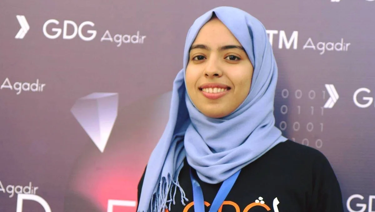 A photo of Hanane, wearing a black GDG  t-shirt and blue head cover and smiling at the camera. In the background is a GDG Agadir DevFest banner.