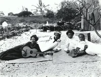 Fotografía en blanco y negro de cuatro mujeres hawaianas que usan vestidos largos y están recostadas sobre lonas en la playa, una de ellas sostiene un ukelele. En el fondo, se pueden observar plantas.