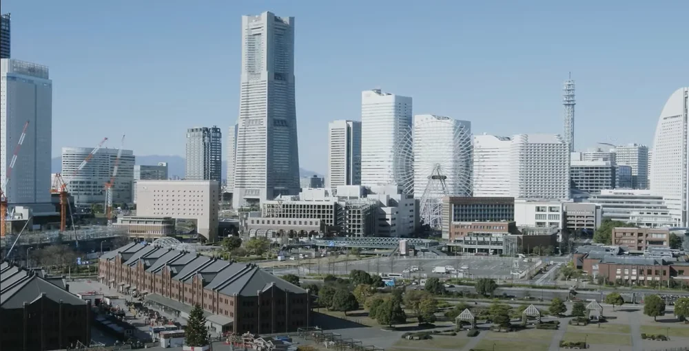 The skyline of of the City of Yokohama in Japan, with low industrial buildings and parkland in the foreground and modern skyscrapers in the background, set against a clear blue sky.