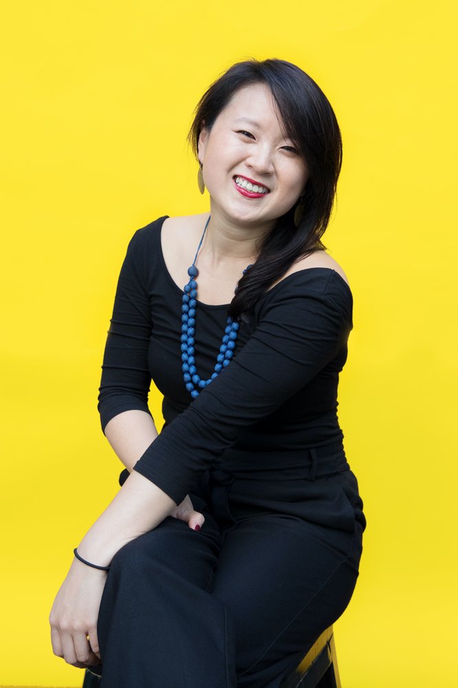 Photo of woman in black clothes and a necklace sitting against a bright yellow back drop.