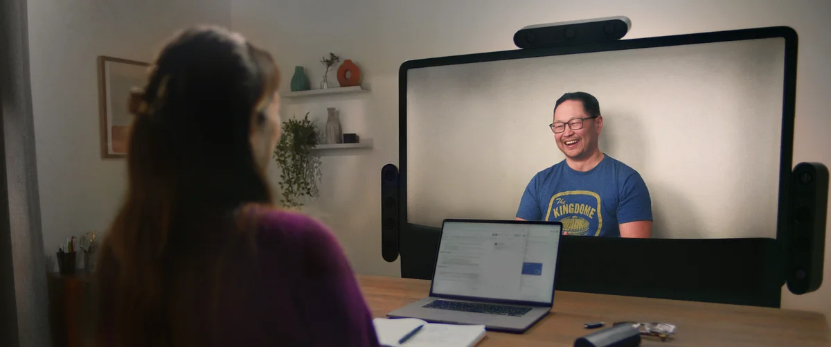 Two people meet on Project Starline. The person on the screen is smiling at the person in the foreground. There is a laptop, notebook and pair of glasses on the table in front of the screen.