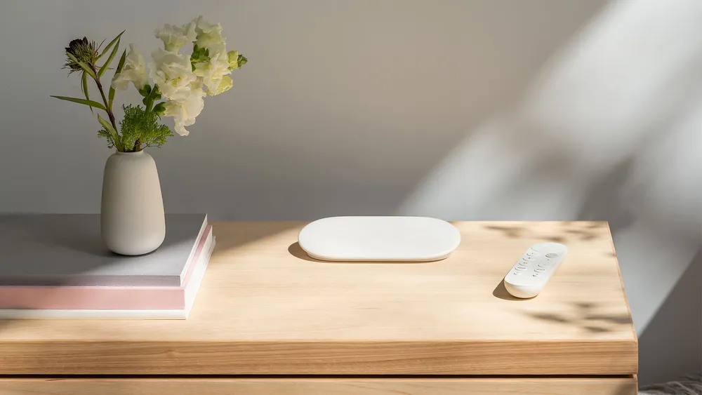 The new Google TV Streamer in Porcelain sits in the middle of a light brown desk. On the right is the new voice remote. On the left is a vase of flowers.