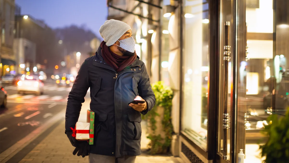 A man wearing a winter coat and a COVID face mask holds two wrapped presents in one hand and his cell phone in the other. His phone is lit up, indicating he was just using it. He is outside of a retail store and looking inside the store window.