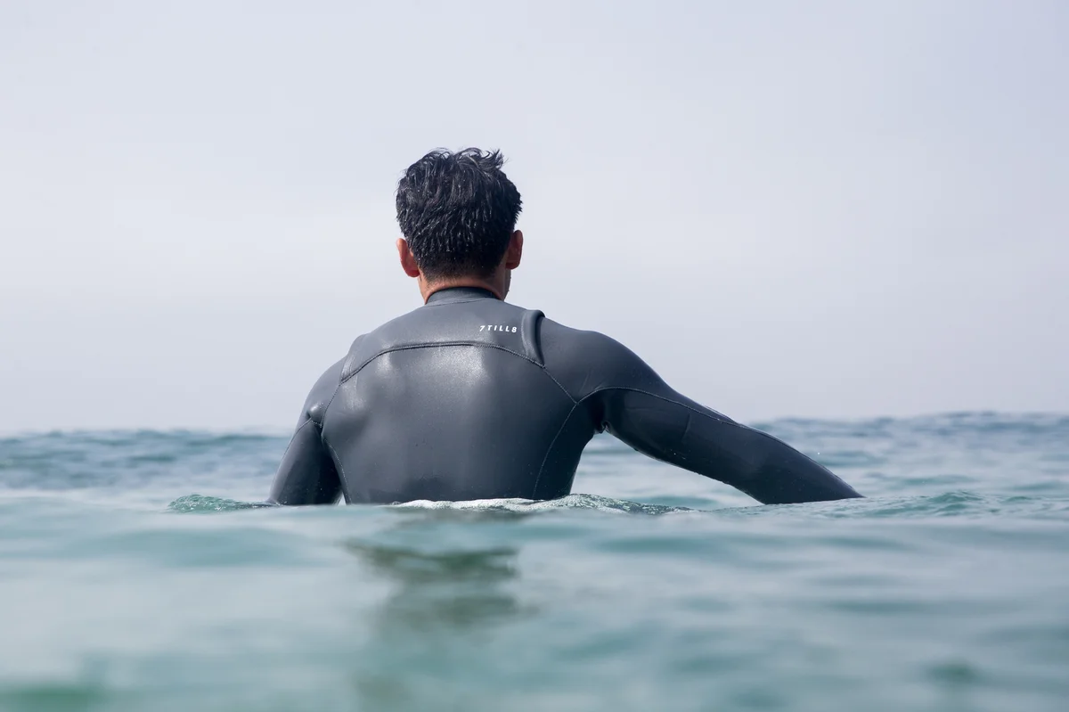 A surfer facing the horizon paddles out in a custom 7TILL8 wetsuit.