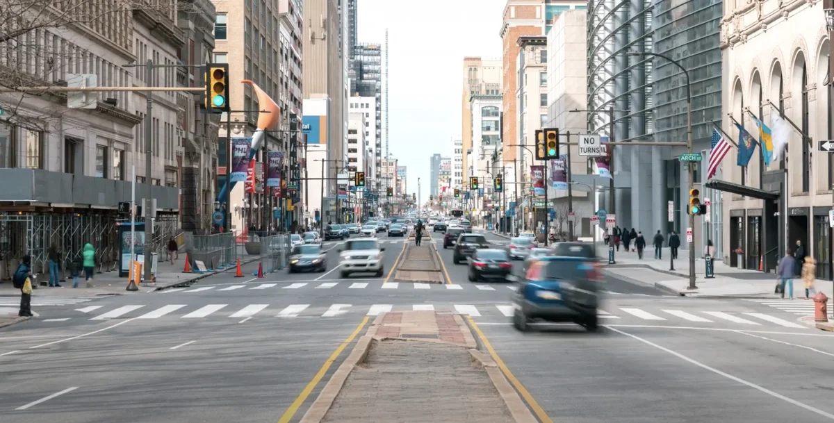 Breite mehrspurige Straße in einer Großstadt, Häuser links und rechts, ein Zebrastreifen, mehrere Autos und Ampeln