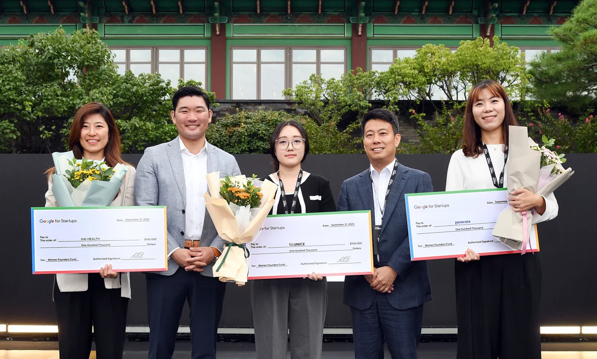 Two men and three women are standing and smiling. The three women are each holding a cheque and bouquet of flowers.