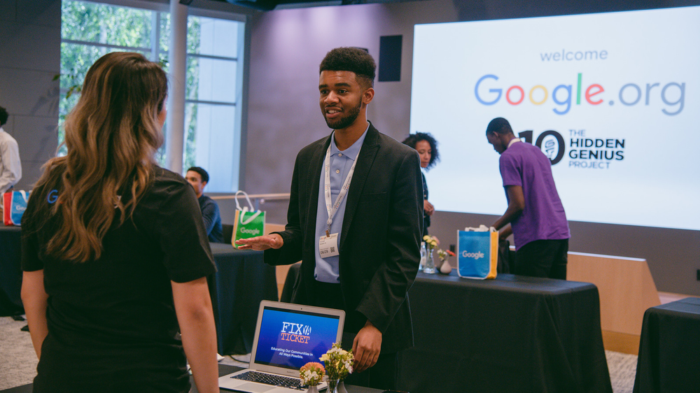
                         
                           A young man in a suit speaks to a woman, who is looking at a laptop. Behind him, a screen shows “Google.org” and “The Hidden Genius Project.”
                         
                       