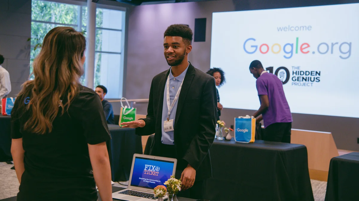 A young man in a suit speaks to a woman, who is looking at a laptop. Behind him, a screen shows “Google.org” and “The Hidden Genius Project.”