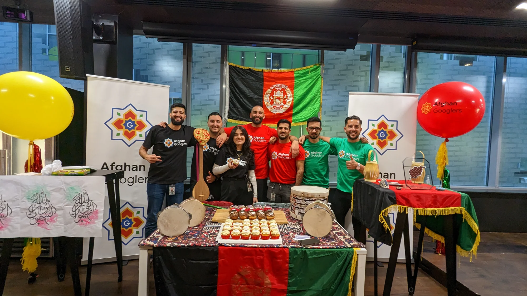 Maryam steht umringt von sechs Kollegen, die alle „Afghan Googlers“-T-Shirts tragen hinter einem Präsentationstisch, auf dem Gebäck sowie Trommelinstrumente liegen. Im Hintergrund ist eine afghanische Flagge zu sehen sowie zwei Roll-ups und Luftballons, auf denen „Afghan Googlers“ steht.