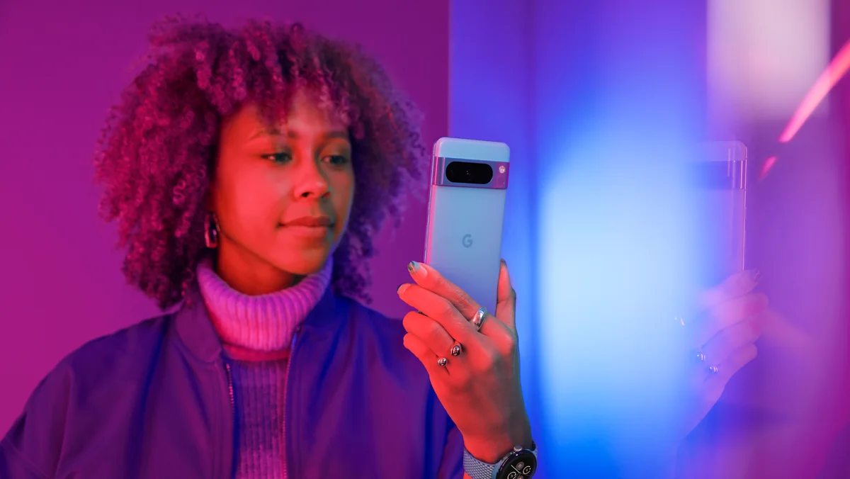 A woman is sitting up in bed. She is holding a Pixel 7 Pro in her left hand and looking at the screen with a slight smile.