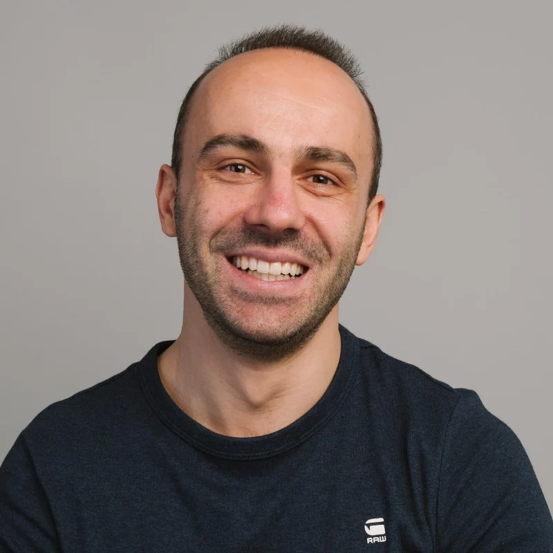 A man wearing a black shirt with short brown hair smiles at the camera in front of a gray background.