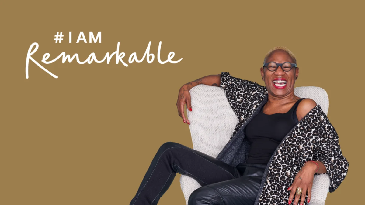 A woman lounges on a white chair and smiles at the camera. #IAmRemarkable text is on the background.