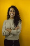 Woman with long dark brown wavy hair stands in front of mustard yellow wall with arms folded, smiling