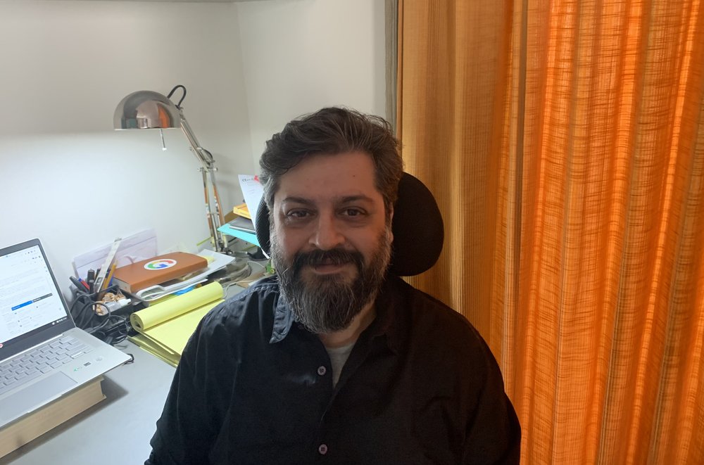 Manav sitting at his desk at home with an orange curtain behind him.