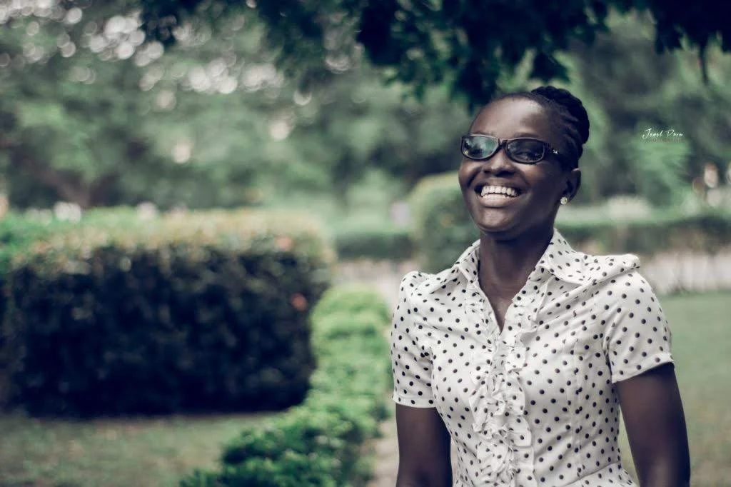 Abigail Annkah is outside against a green garden backdrop wearing a white and black polka dot shirt and sunglasses and smiling warmly
