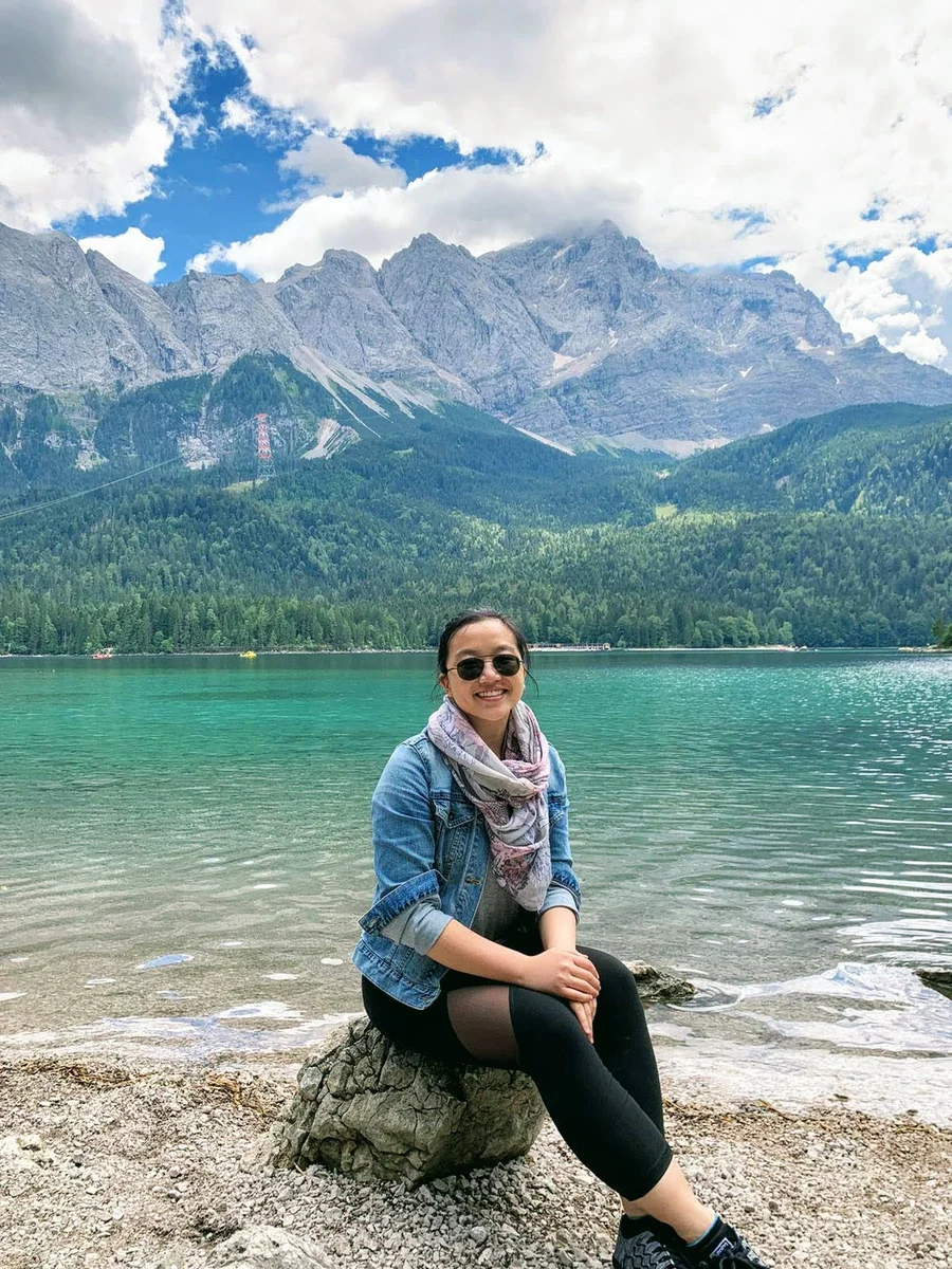 Marisa Chow sitzt auf einem Stein vor dem Eibsee in Bayern, im Hintergrund ist die Zugspitze zu sehen