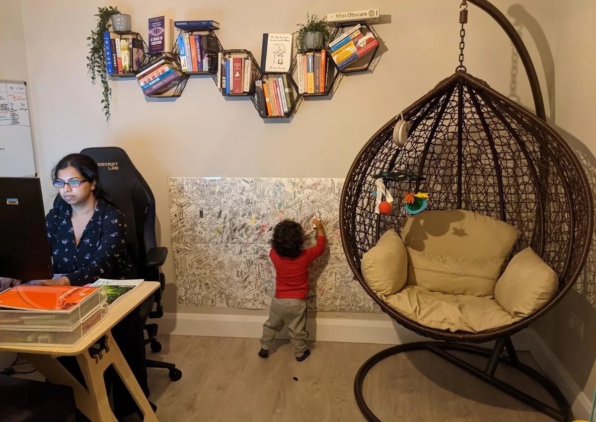 Krithika sitting at a desk working on a monitor. Behind her, her two year old child draws on paper stuck to the wall. Bookshelves and a swinging chair also decorate the room.