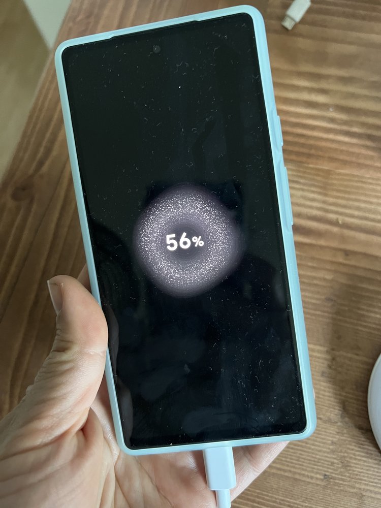 A photograph of a hand holding a plugged in Pixel 6a. The screen has a light-up effect around a "56%" notification.