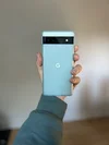 A photograph of a hand holding a Pixel 6a, with the back of the phone facing the camera. The phone is in a blue-green semi-translucent case. There is a "G" Google icon on it.