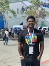 Saad wearing a black shirt with a multicolored globe image on it. Behind him is a white building, trees, and people walking around.