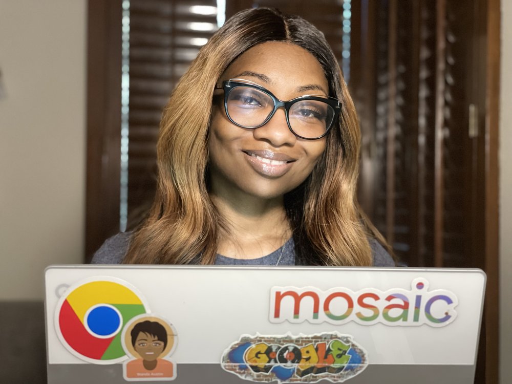 Shameeka is wearing black glasses and a gray shirt, and smiling at the camera. We can see her from the shoulders up, as she is sitting in front of a laptop covered in colorful stickers. Behind Shameeka is a window with brown shades.