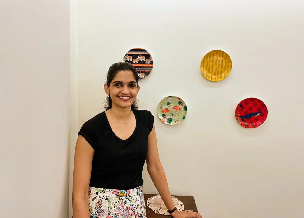 Author Sneha Srinivasan smiling, in a black top and white patterned skirt, leaning against a table to her left, with colourful ceramic plates on the white wall behind her.
