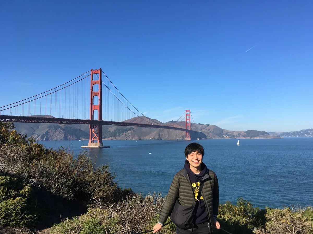 Inho posing with a smile in front of the Golden Gate Bridge. In the background is a red bridge stretching across a wide body of blue water, with sloping hills on the far side of the bridge. In the foreground are some green bushes.
