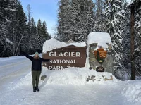 Anne is wearing winter clothes and standing on a snowy road next to a sign that says "Glacier National Park."