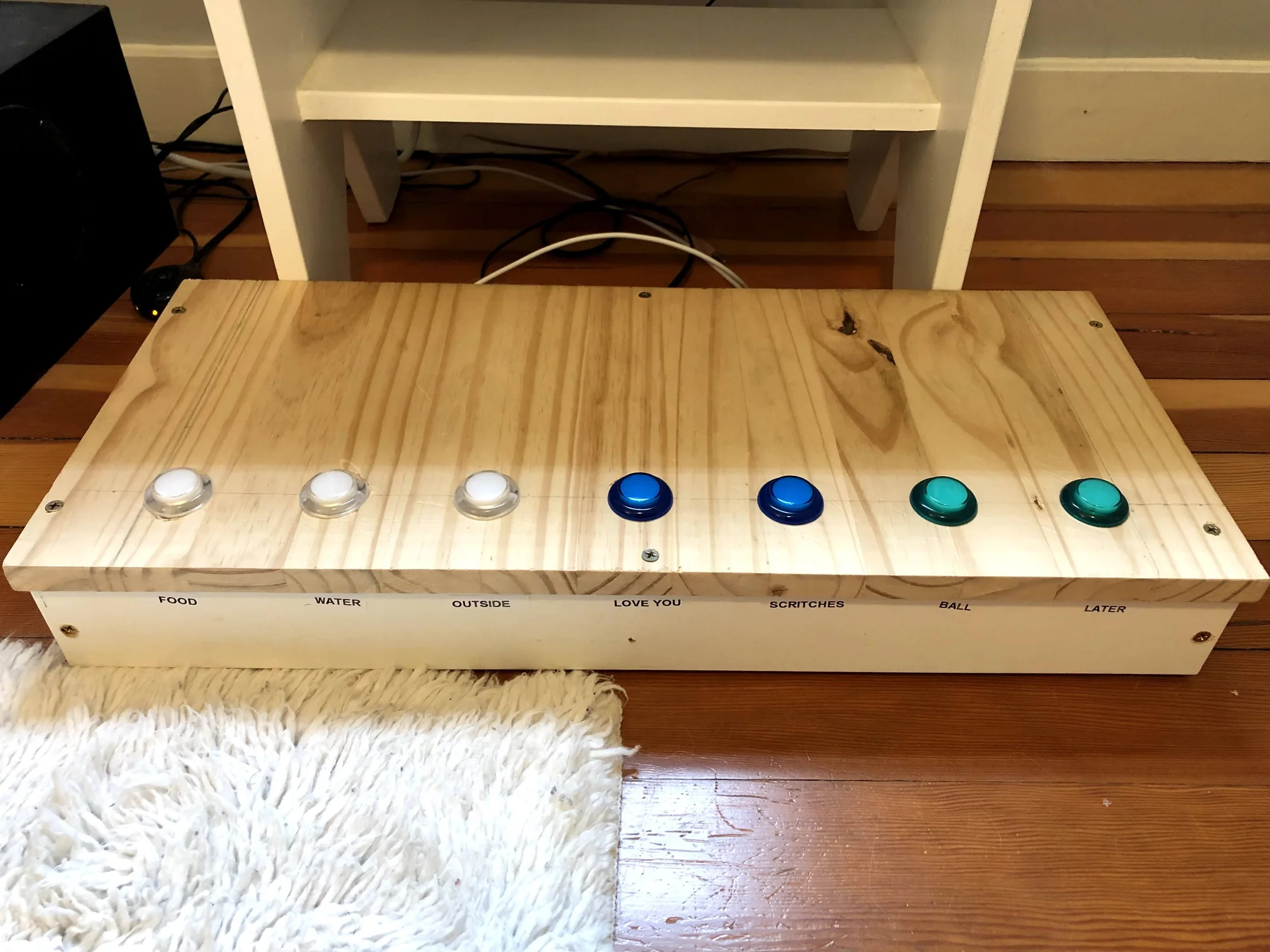 A rectangular wooden board with three white buttons, two blue buttons and two green buttons. Underneath each button is “food,” “water,” “outside,” “love you,” “scritches,” “ball” and “later.”